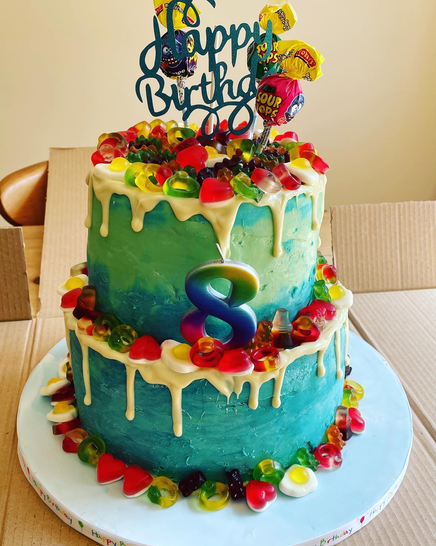 Picture depicts two tier sponge cake with choice of filling and is decorated with white chocolate ganache (coloured blue) with contrasting white chocolate drip, topped with sweets.